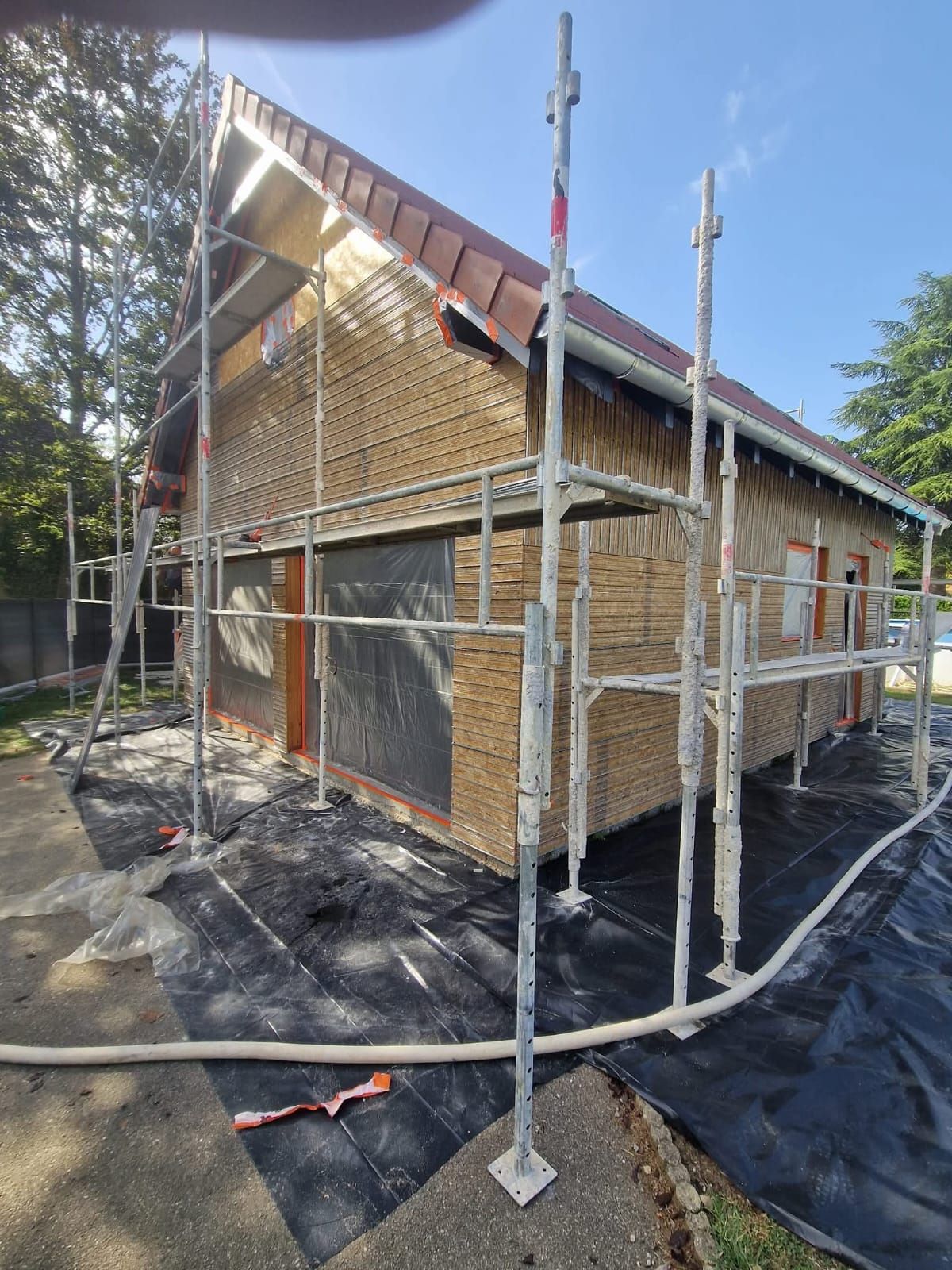 Bâtiment en construction, des échafaudages entourent une maison au bardage en bois et au toit de tuiles brunes.