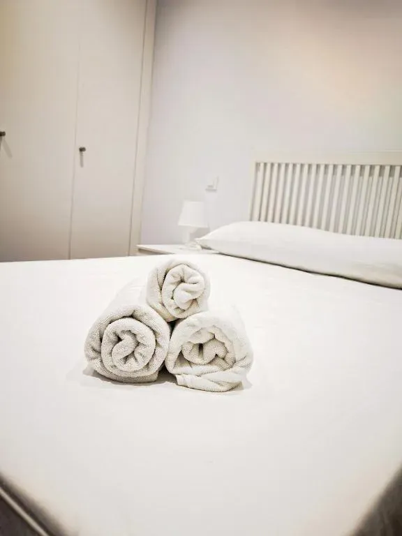 Cama blanca con toallas enrolladas, mesita de noche y armario en una habitación luminosa.