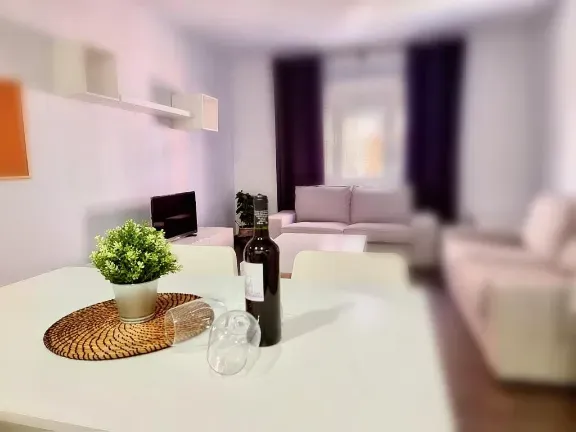 Mesa puesta para comer con vino, copas y una planta en una moderna sala de estar con sofá y cortinas.