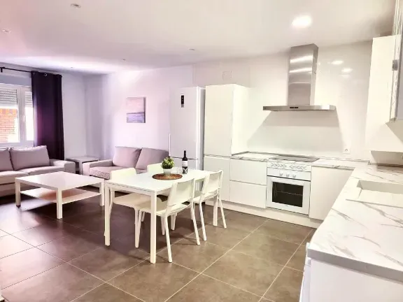 Espacio diáfano con cocina, mesa de comedor y sofá. Armarios y electrodomésticos blancos.