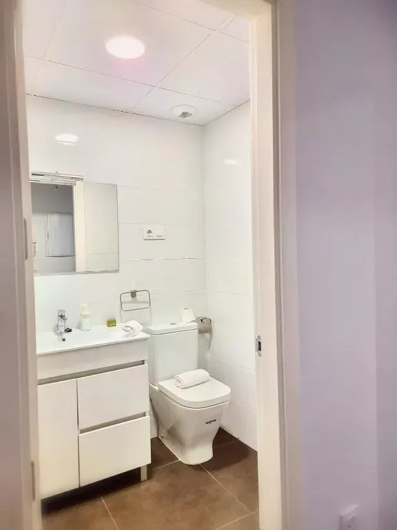 Un baño pequeño y blanco con inodoro, lavabo y espejo, visto desde la puerta.