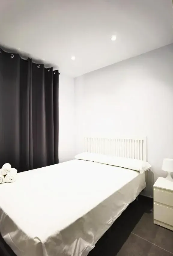 Dormitorio de paredes blancas con cama, mesita de noche y cortinas oscuras.