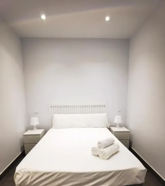 Dormitorio blanco con cama, mesitas de noche, lámparas y toallas enrolladas.
