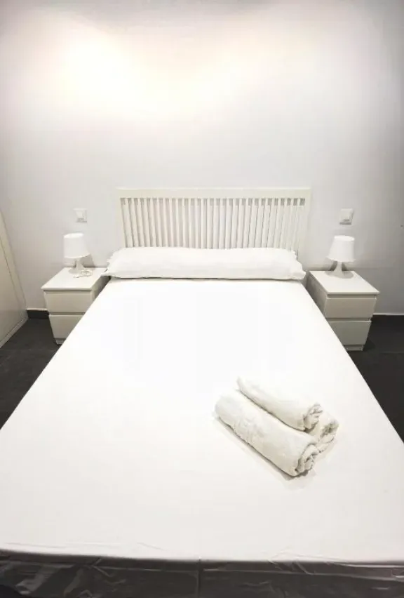 Dormitorio de paredes blancas con cama grande, dos mesitas de noche con lámparas y toallas enrolladas sobre la cama.