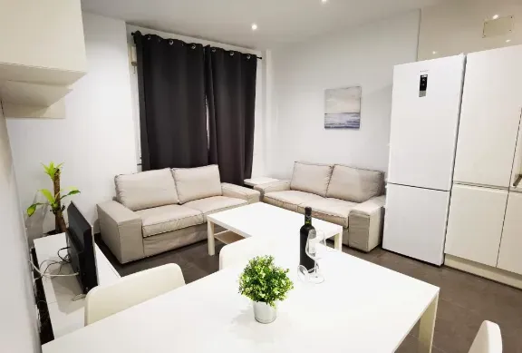 Un espacio de estar luminoso con dos sofás beige, una mesa blanca y un refrigerador.