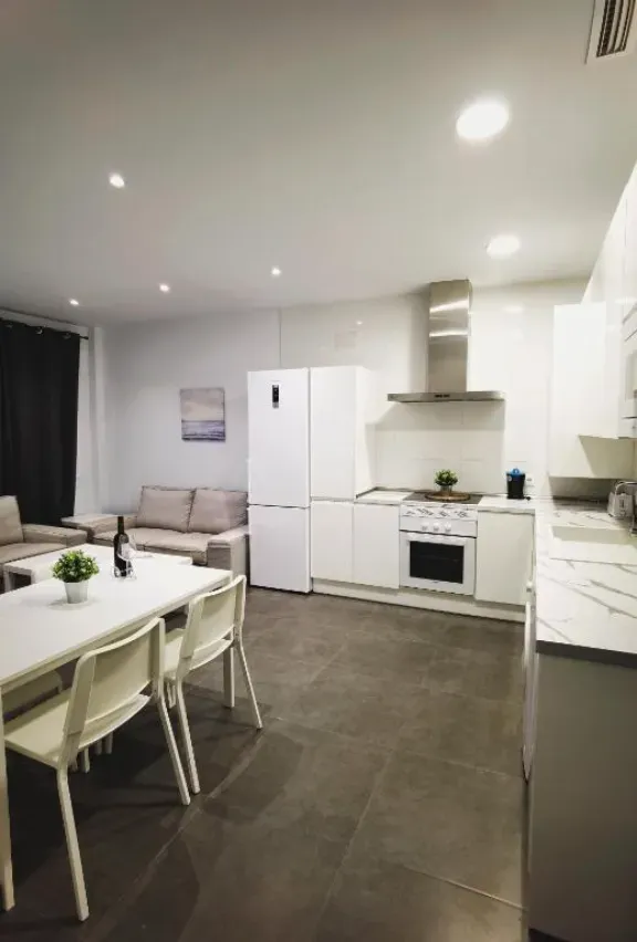 Cocina y comedor modernos y luminosos. Armarios, electrodomésticos, mesa y sillas blancos. Suelo y sofá gris oscuro.