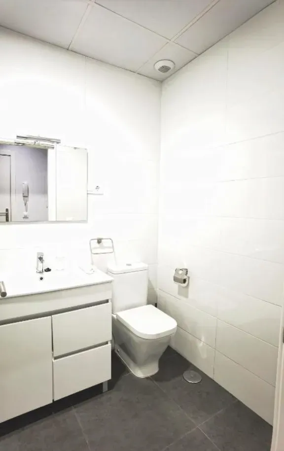 Baño blanco con inodoro, lavabo, espejo y suelo de baldosas grises.