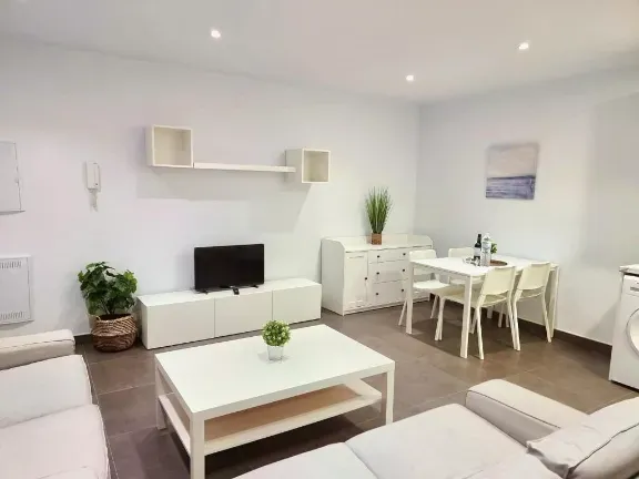 Sala de estar luminosa y moderna con muebles blancos, TV y mesa de comedor.