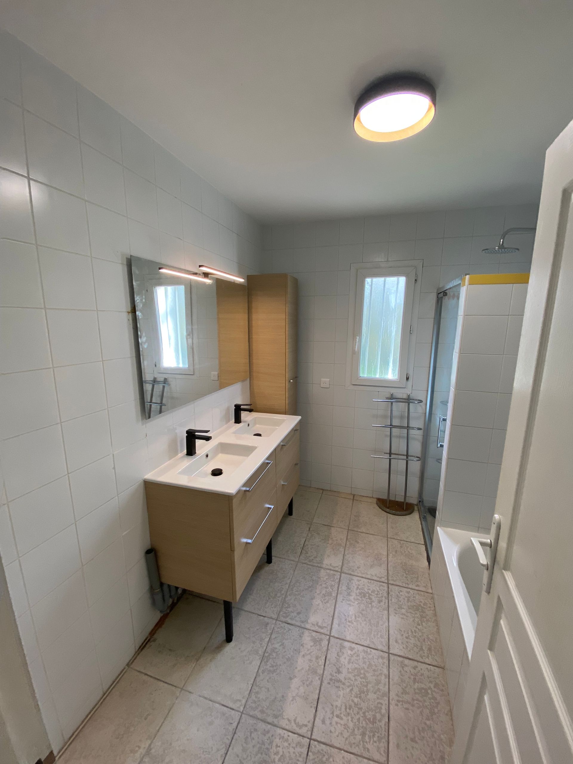 Petite salle de bains moderne avec murs carrelés blancs, meuble-lavabo beige, miroir, fenêtre et cabine de douche.