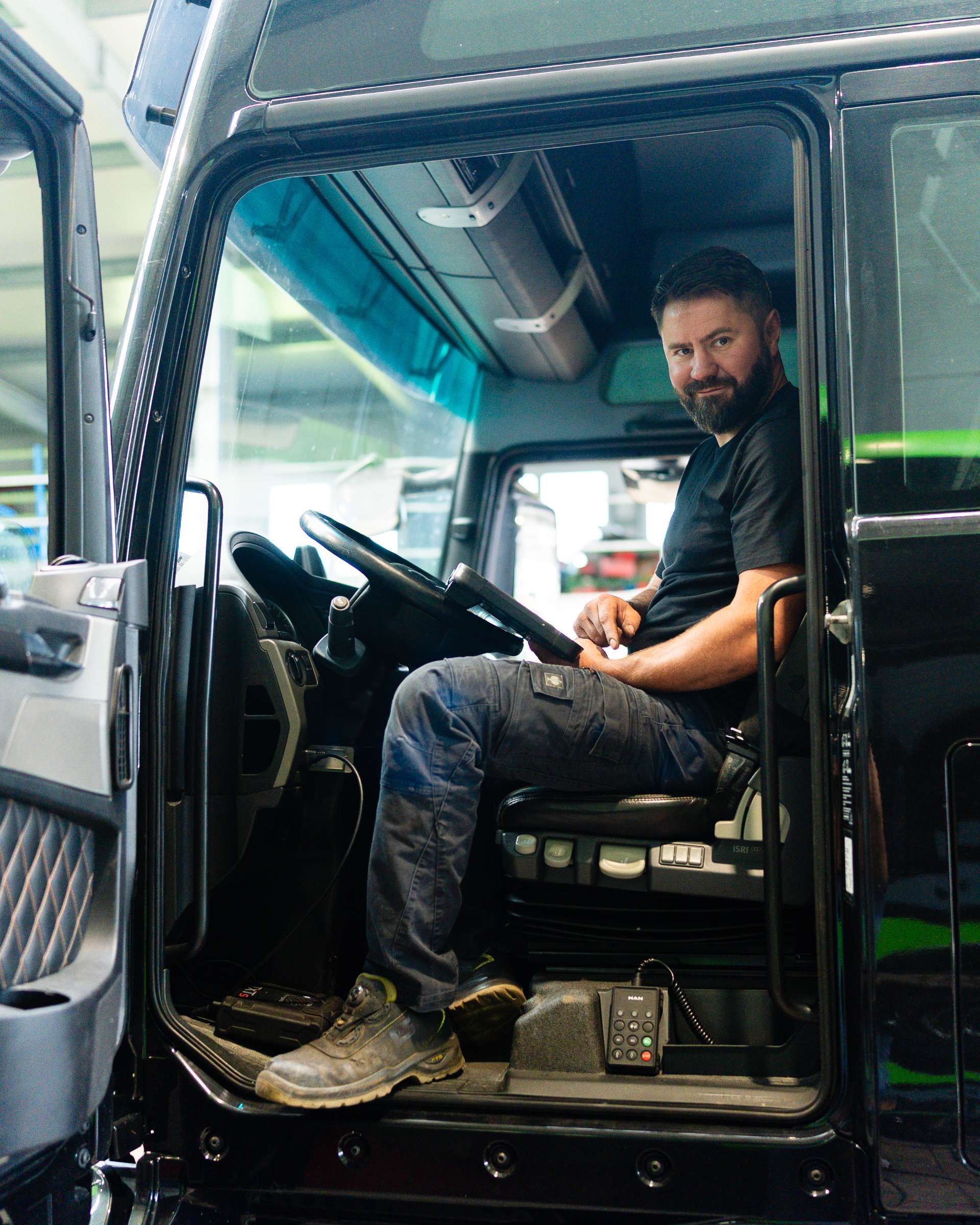 Ein Mann sitzt auf dem Fahrersitz eines schwarzen Lastwagens, die Tür ist offen. Er lächelt und schaut in die Kamera.