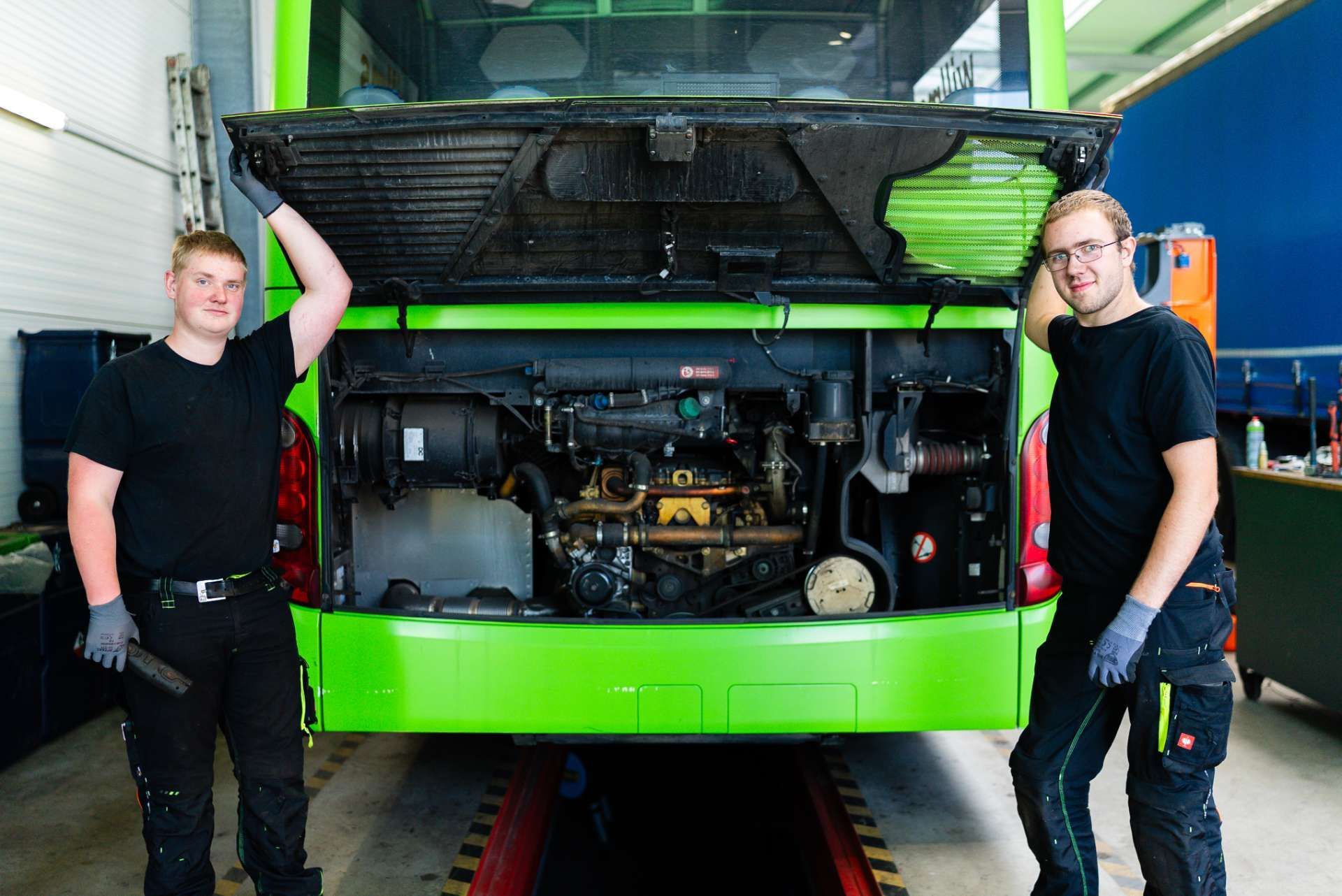 Zwei Mechaniker in schwarzen Hemden stehen neben einem Bus mit offenem Motorraum und grüner Außenlackierung.
