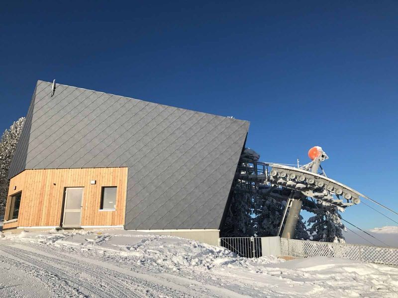 Verschneite Skiliftstation mit grauem Dach und Holzelementen, klarer blauer Himmel.
