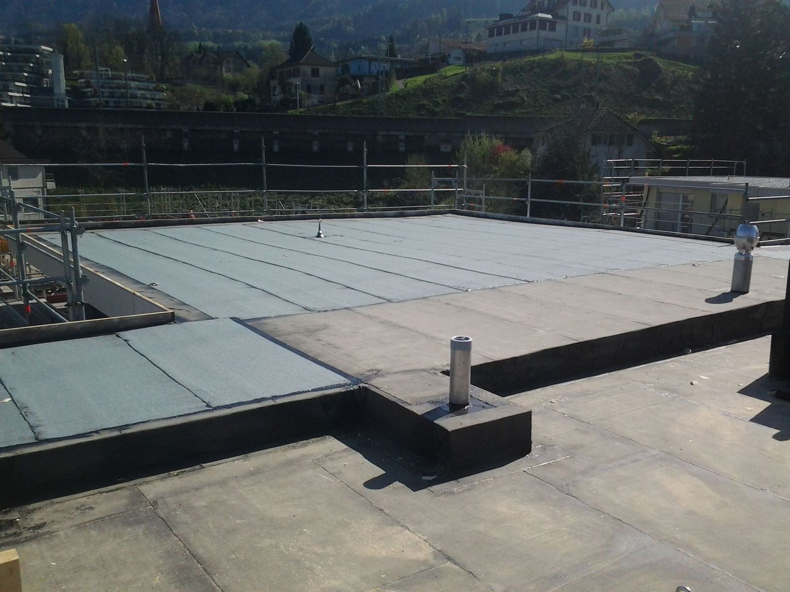 Dachterrasse mit schwarzem Flachdach, hellgrauen quadratischen Elementen, Geländer und einer Person am Rand. Im Hintergrund Gebäude und Bäume.