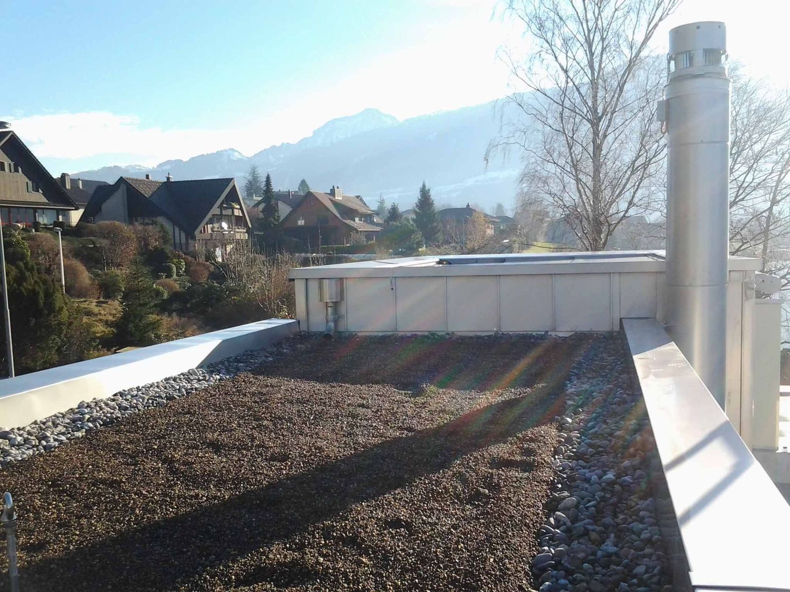 Dachgarten mit Kieselsteinen, Betonkonstruktion und Schornstein. Im Hintergrund Häuser und Berge unter blauem Himmel.