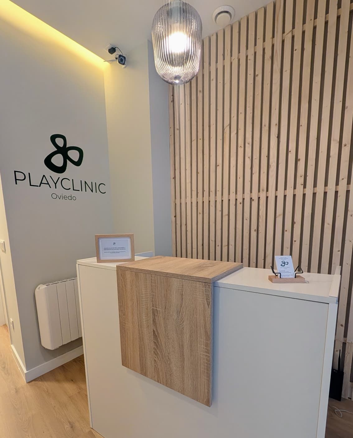 Zona de recepción de una clínica de fisioterapia, logotipo “Playfiso Oviedo”, pared de paneles de madera, silla, mesa pequeña.