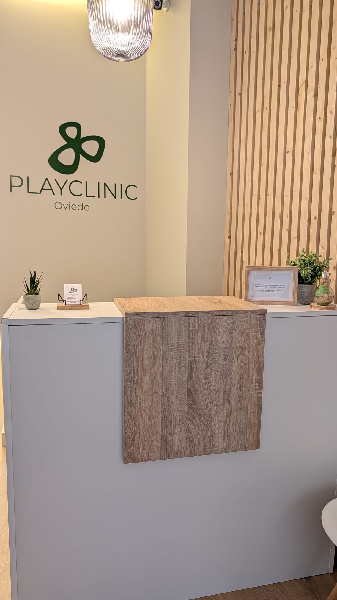 Zona de recepción de una clínica de fisioterapia, logotipo “Playfiso Oviedo”, pared de paneles de madera, silla, mesa pequeña.