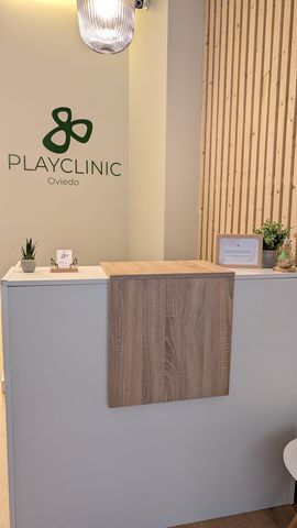Zona de recepción de una clínica de fisioterapia, logotipo “Playfiso Oviedo”, pared de paneles de madera, silla, mesa pequeña.