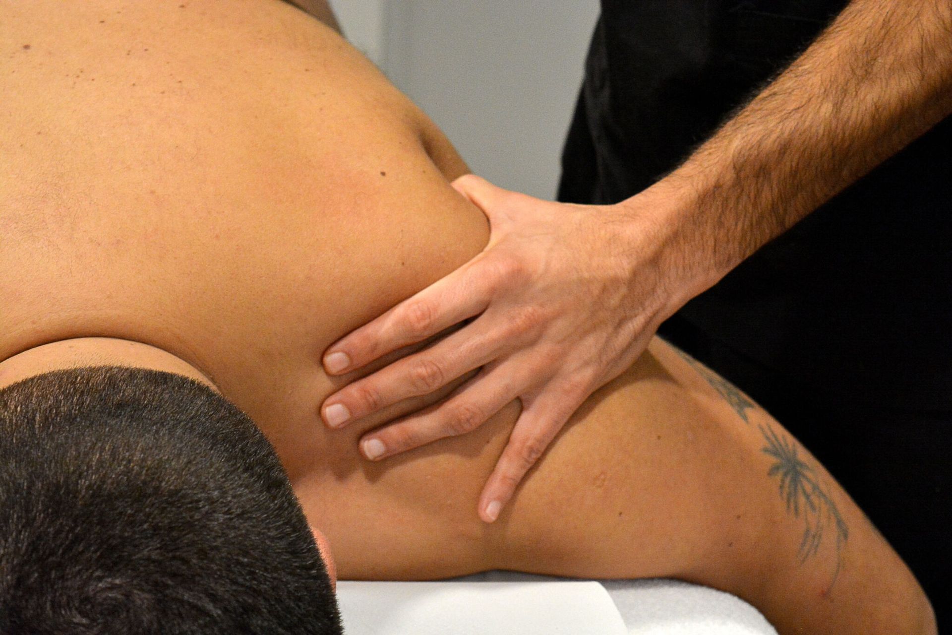 Persona recibiendo un masaje de hombros, torso expuesto, brazo con tatuaje, configuración blanca.