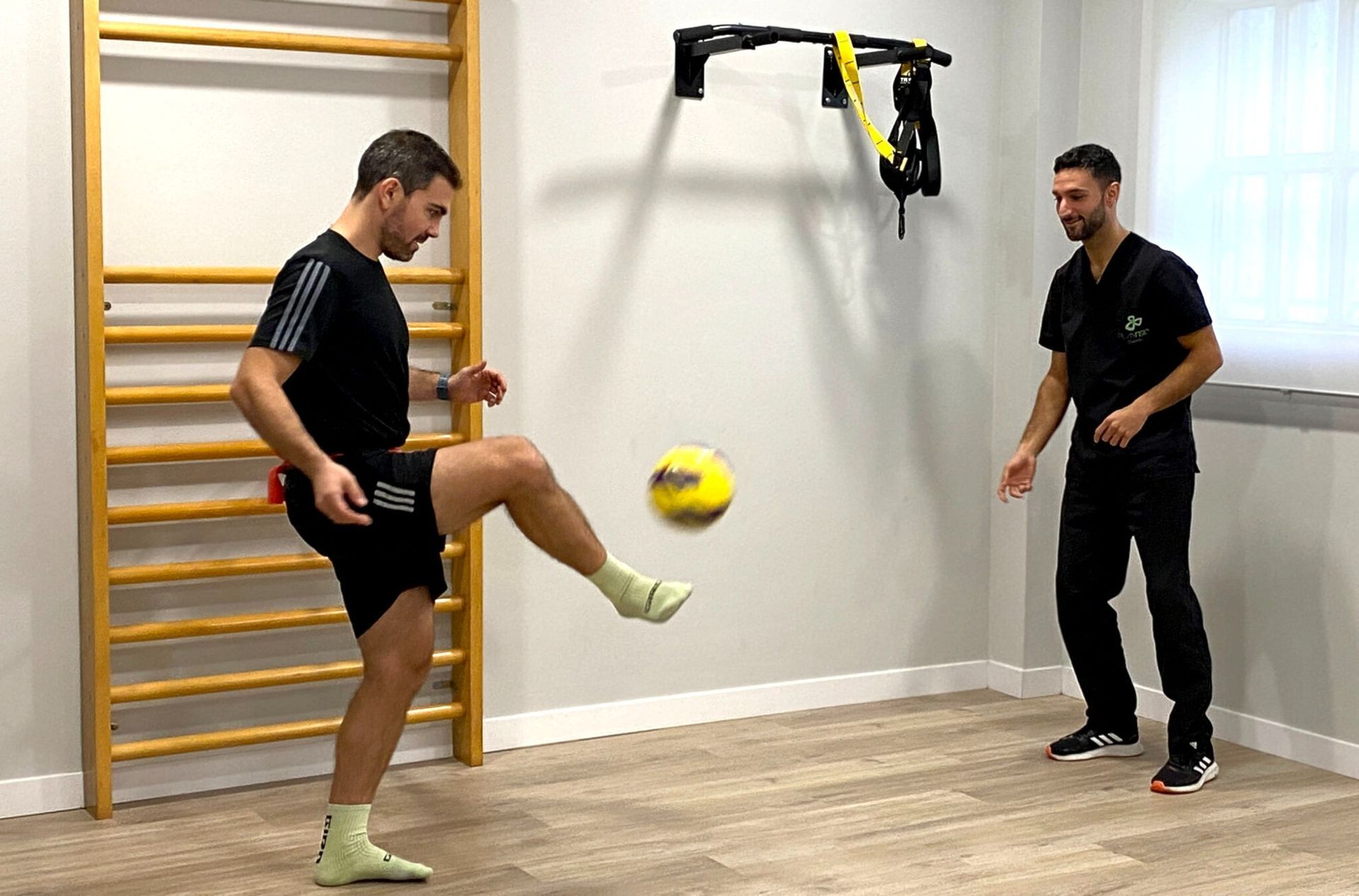 Un hombre patea un balón de fútbol en una sala de terapia, mientras otro hombre observa. Calcetines blancos, pantalones cortos negros, paredes beige.