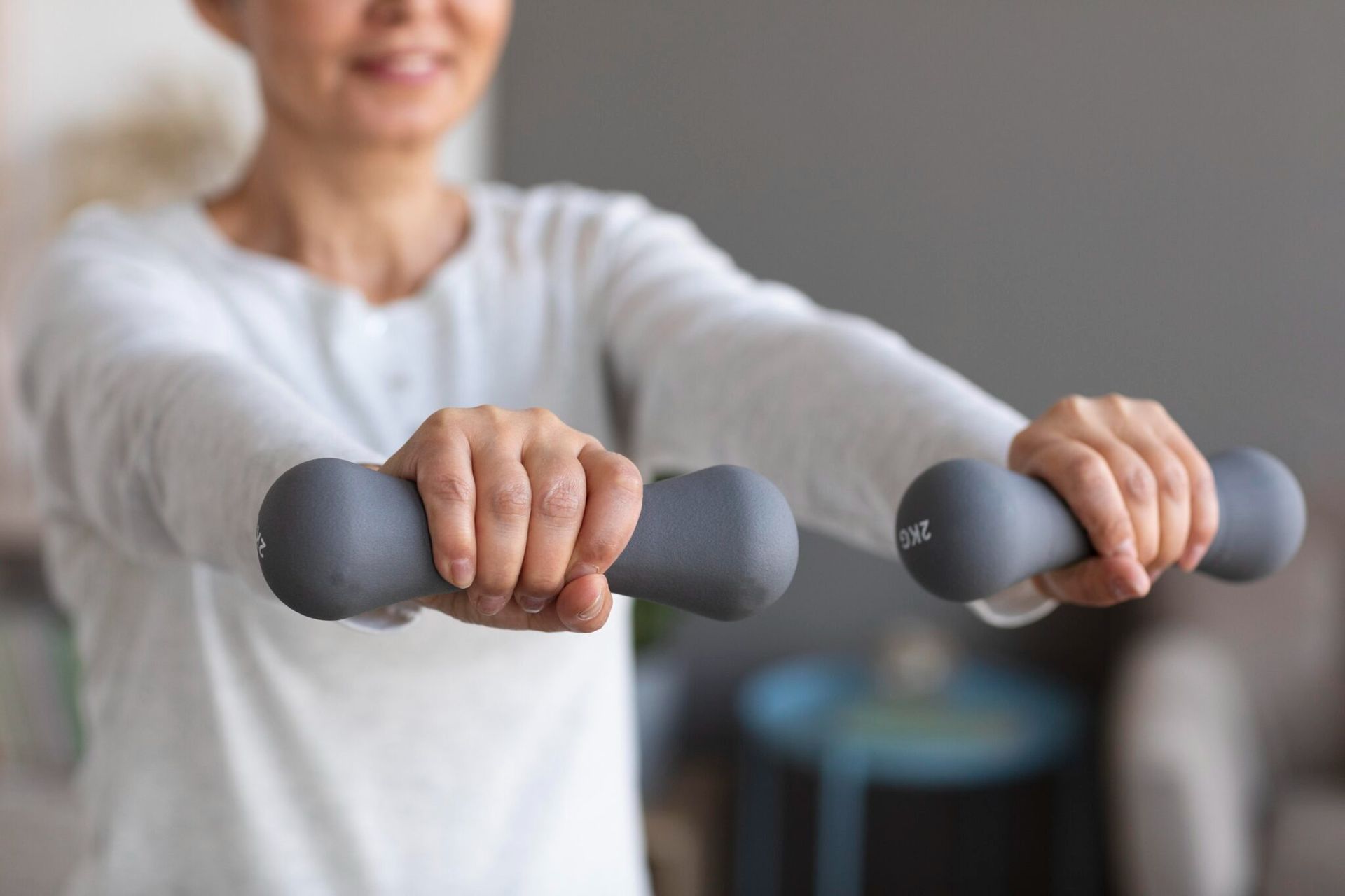 Mujer sosteniendo mancuernas, con los brazos extendidos, haciendo ejercicio en el interior.