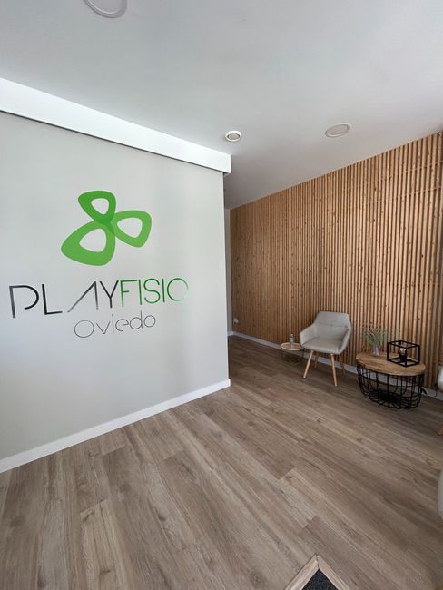 Interior de la clínica Playfisio; logotipo verde en la pared, paneles de madera, silla y mesa.