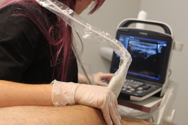Persona con guantes utilizando una máquina de ultrasonido en la pierna de un paciente; examen médico en curso.