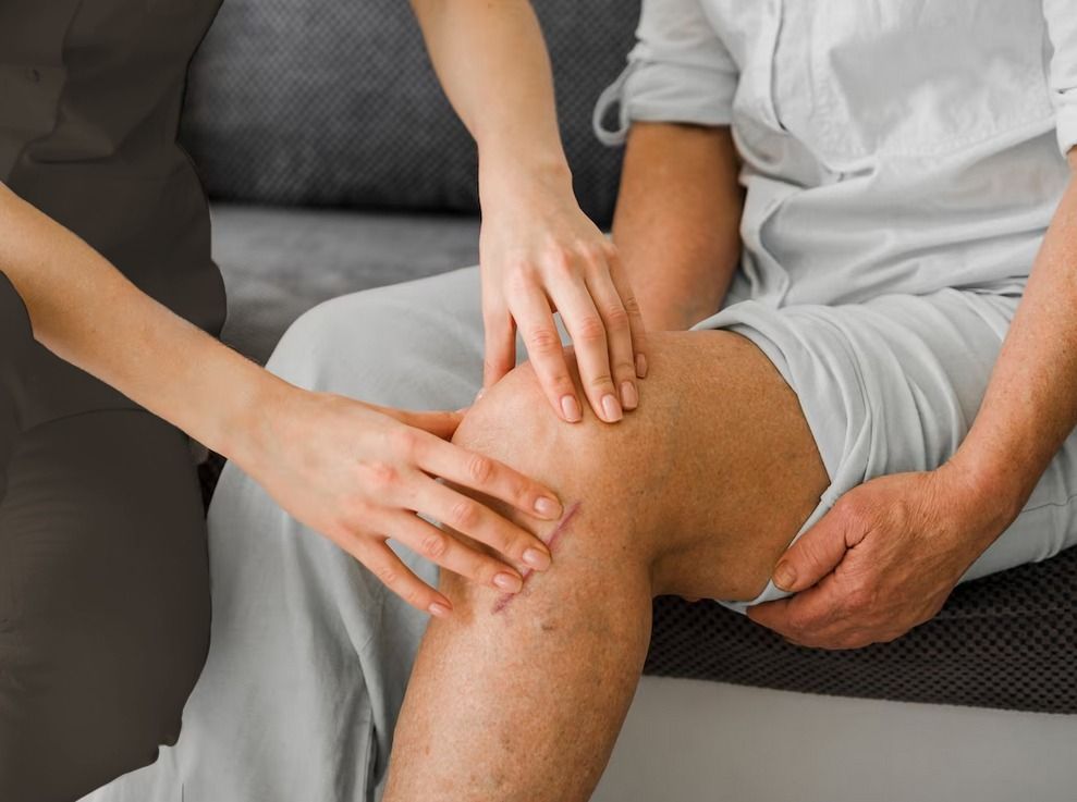 Un profesional médico examina la rodilla de un hombre mayor. Unas manos la tocan suavemente; el hombre está sentado en un sofá gris.