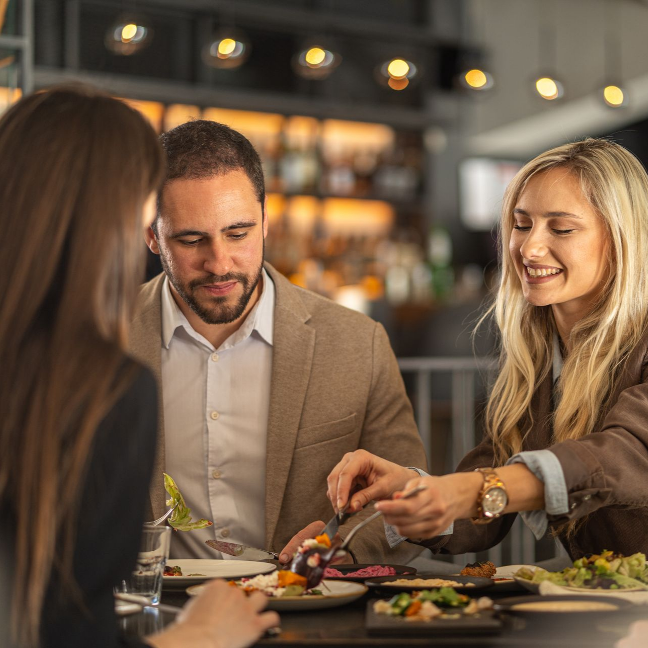 Trois personnes dînent ensemble au restaurant, partageant un repas et souriant par-dessus leurs assiettes et leurs boissons.