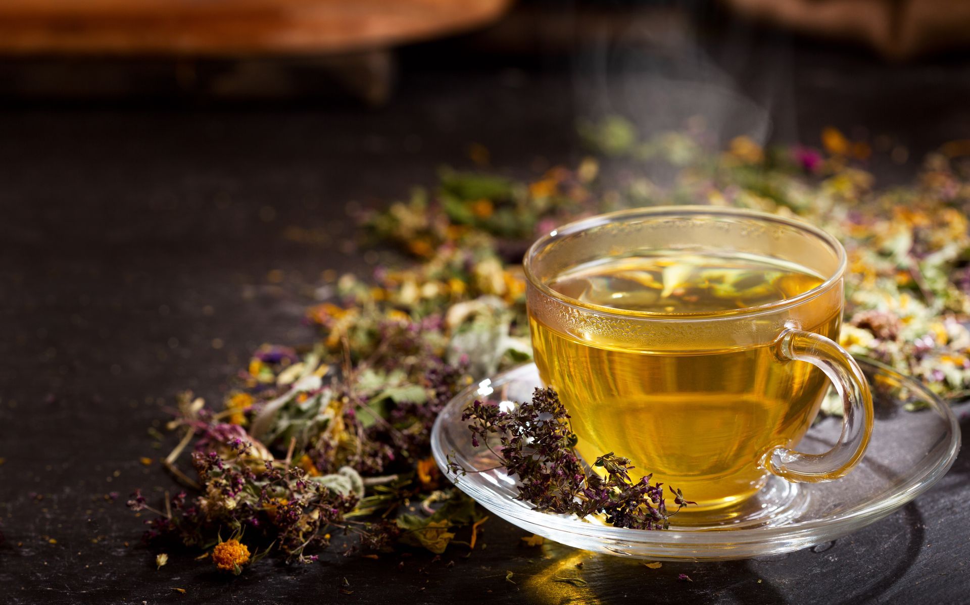 Tasse de thé ambré en verre sur une soucoupe, à côté d'herbes séchées en vrac sur une table sombre.
