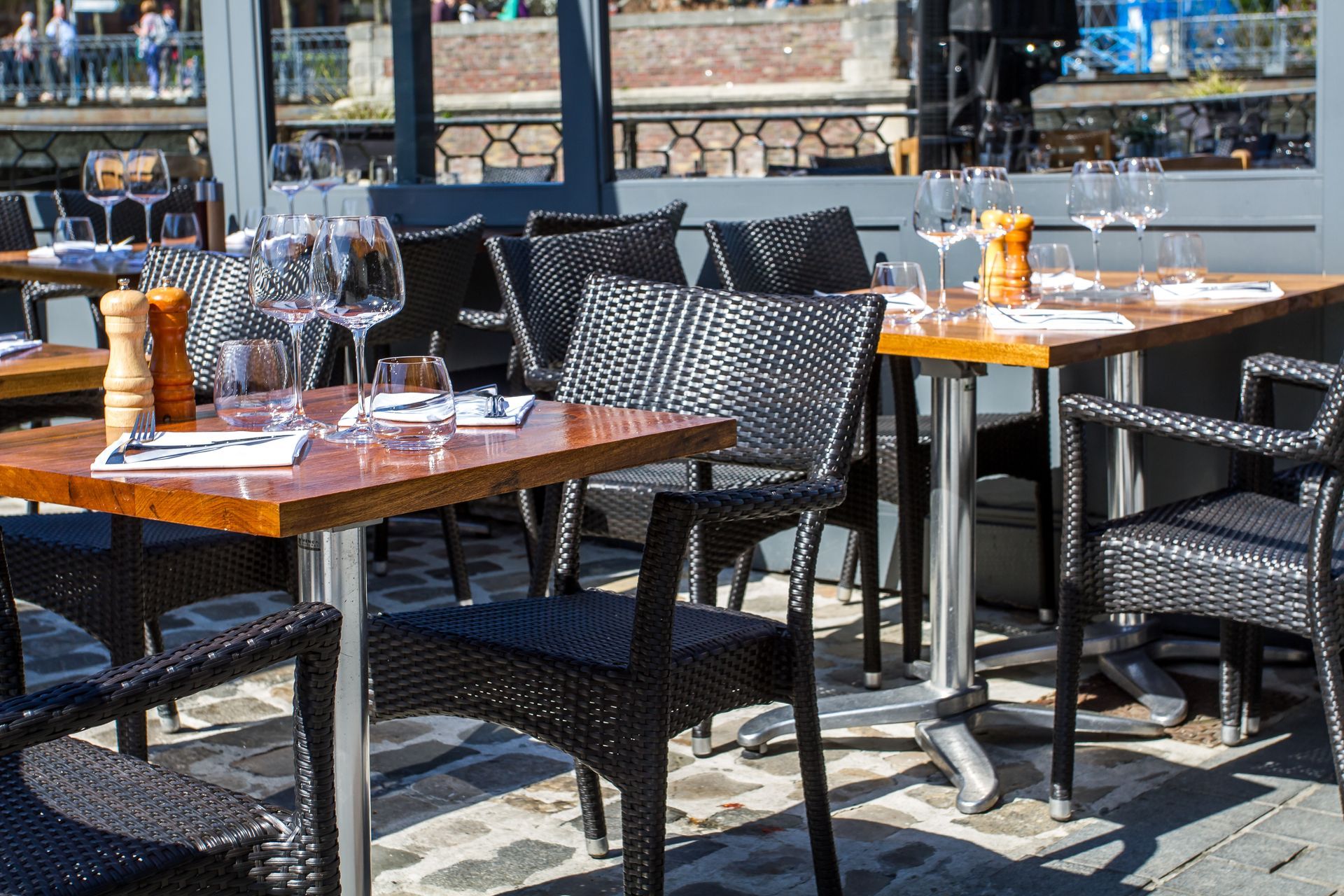 Terrasse ensoleillée avec tables de café en plein air, chaises en osier noir et verres à vin.