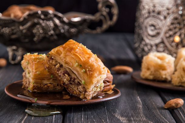 Des baklavas sur une table sombre, avec des pistaches et de l'argenterie ornée en arrière-plan.