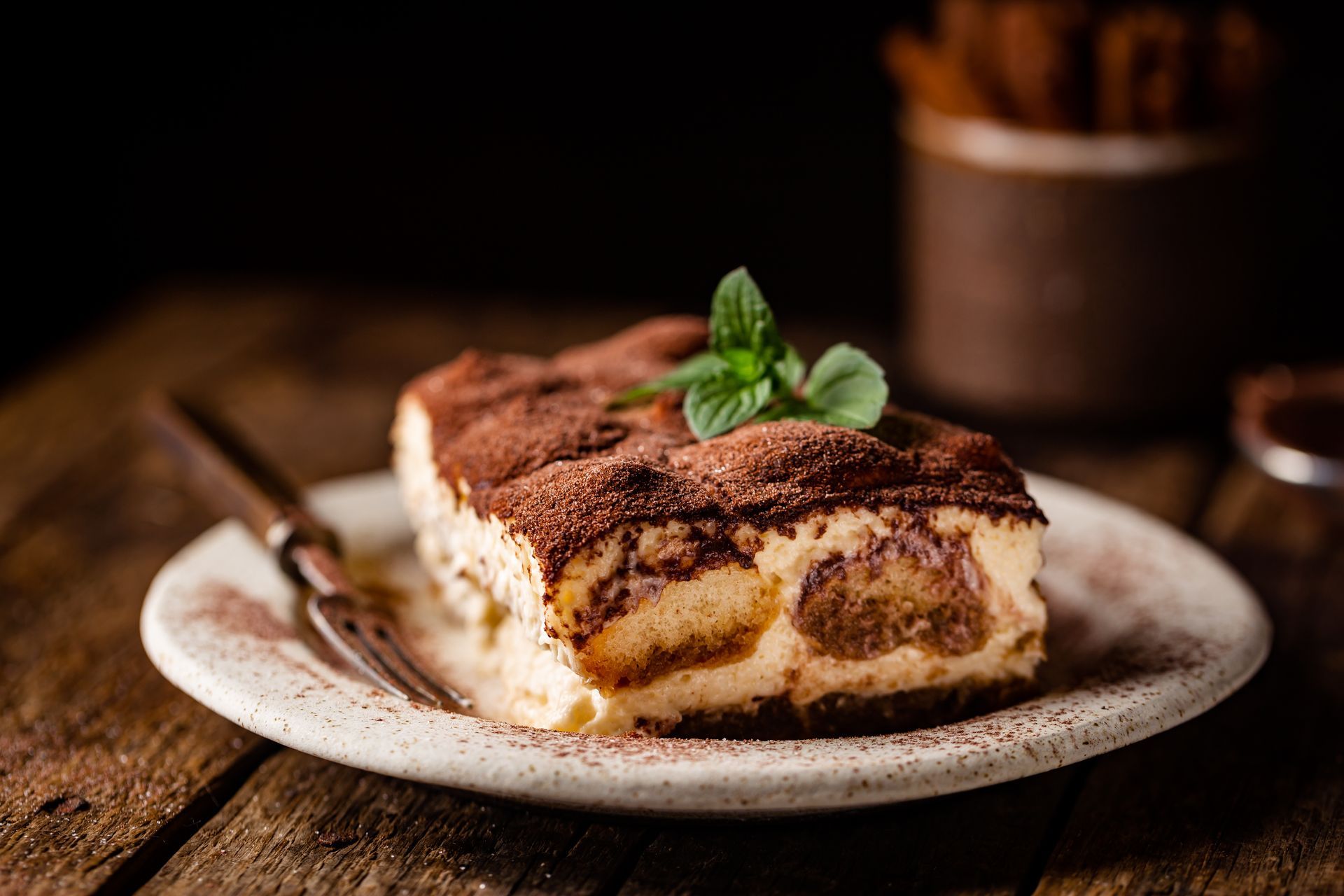 Une part de tiramisu sur une assiette, nappée de cacao et de menthe, sur une table rustique en bois.