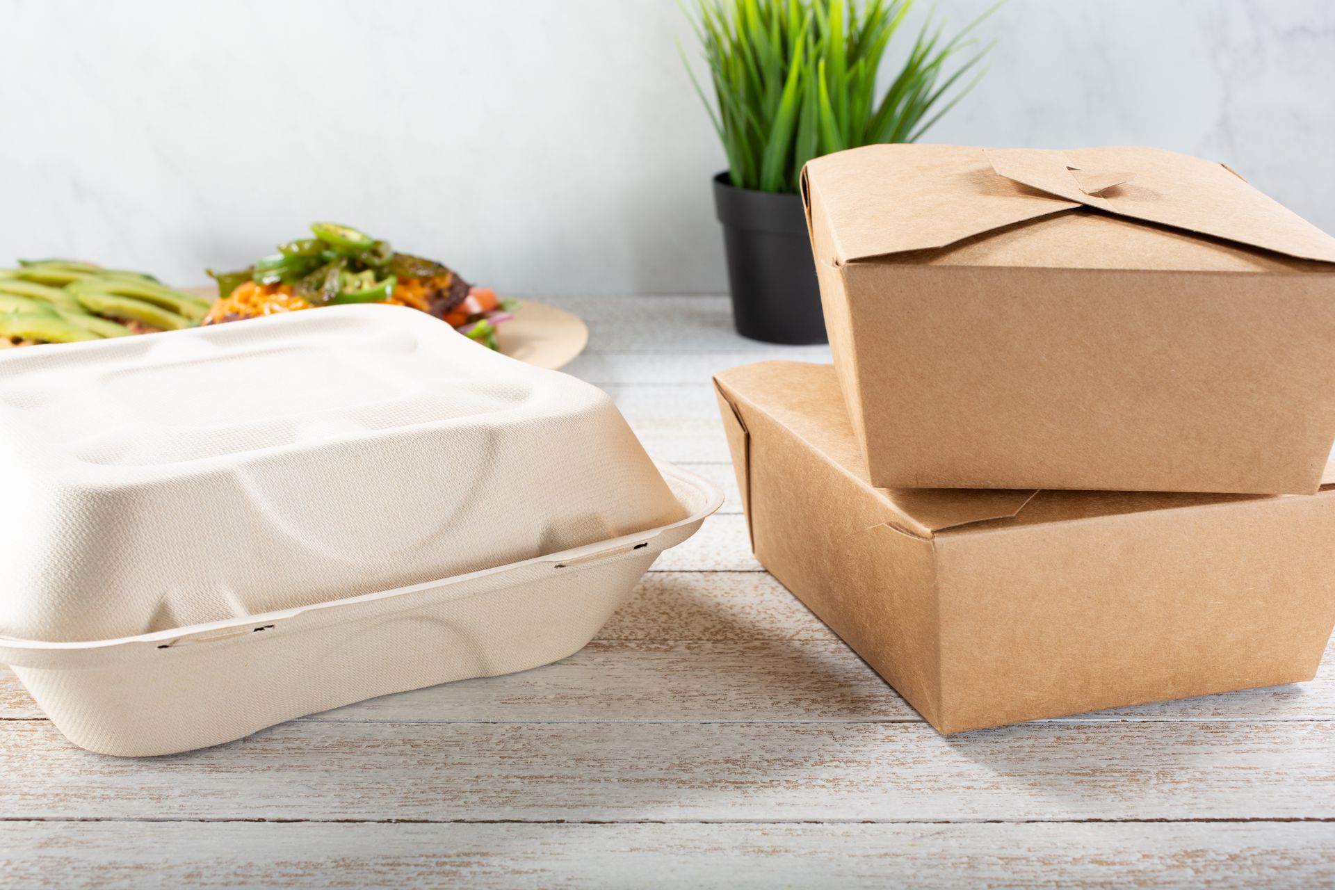 Des boîtes de plats à emporter empilées sur une table, avec de la salade et une petite plante en pot en arrière-plan.