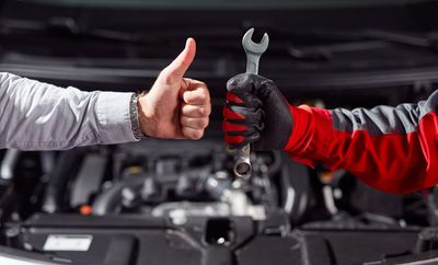 Hände von zwei Personen vor der Motorhaube eines Autos. Eine Person hält ihren Daumen hoch, die andere Person hält Werkzeug in der Hand.