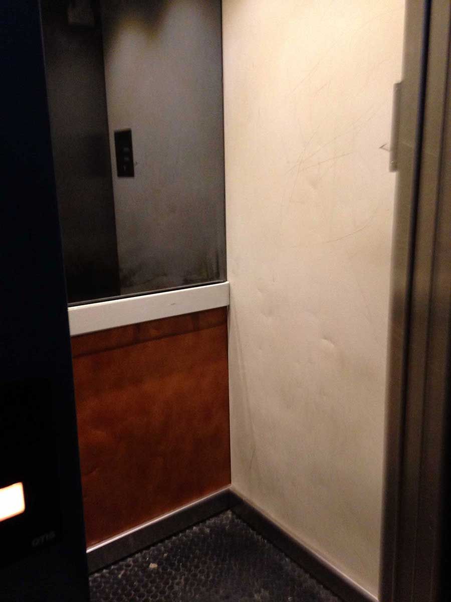 Interior del ascensor: la puerta abierta revela paredes de metal oscuro, paneles marrones, piso negro y panel de control.