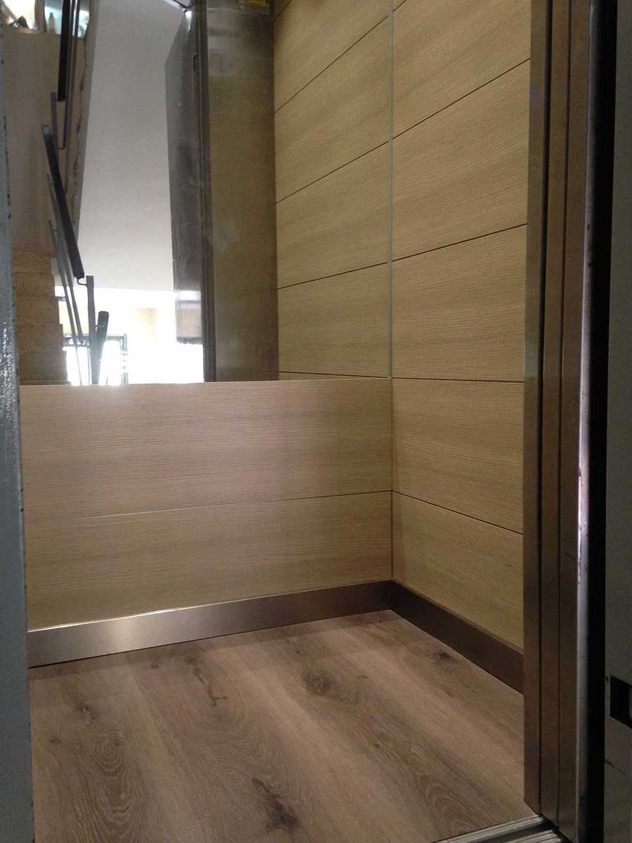 Dentro de un ascensor, paredes revestidas de madera, pared trasera con espejo, suelo de madera de color claro, puerta abierta.