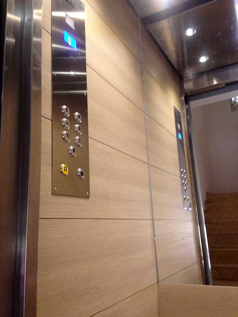 Interior de un ascensor con botones e indicadores de planta. Revestimiento de madera en las paredes, molduras metálicas y escaleras visibles.