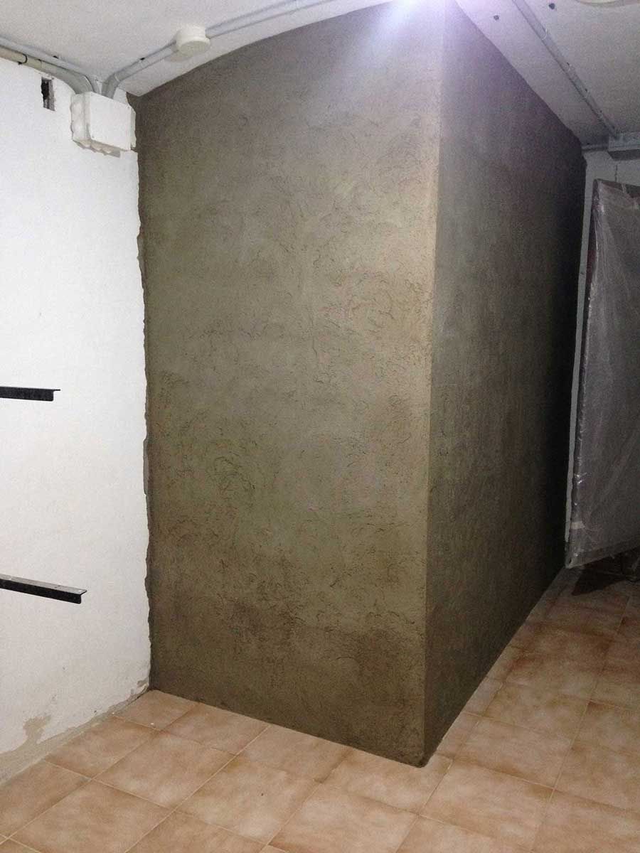 Esquina de una habitación con pared de hormigón, suelo de baldosas y tuberías a la vista.