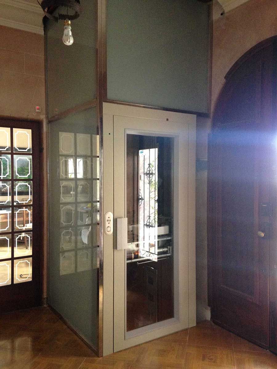 Ascensor en un edificio, de cristal y metal con paneles esmerilados y transparentes. Tonos blancos y marrones.