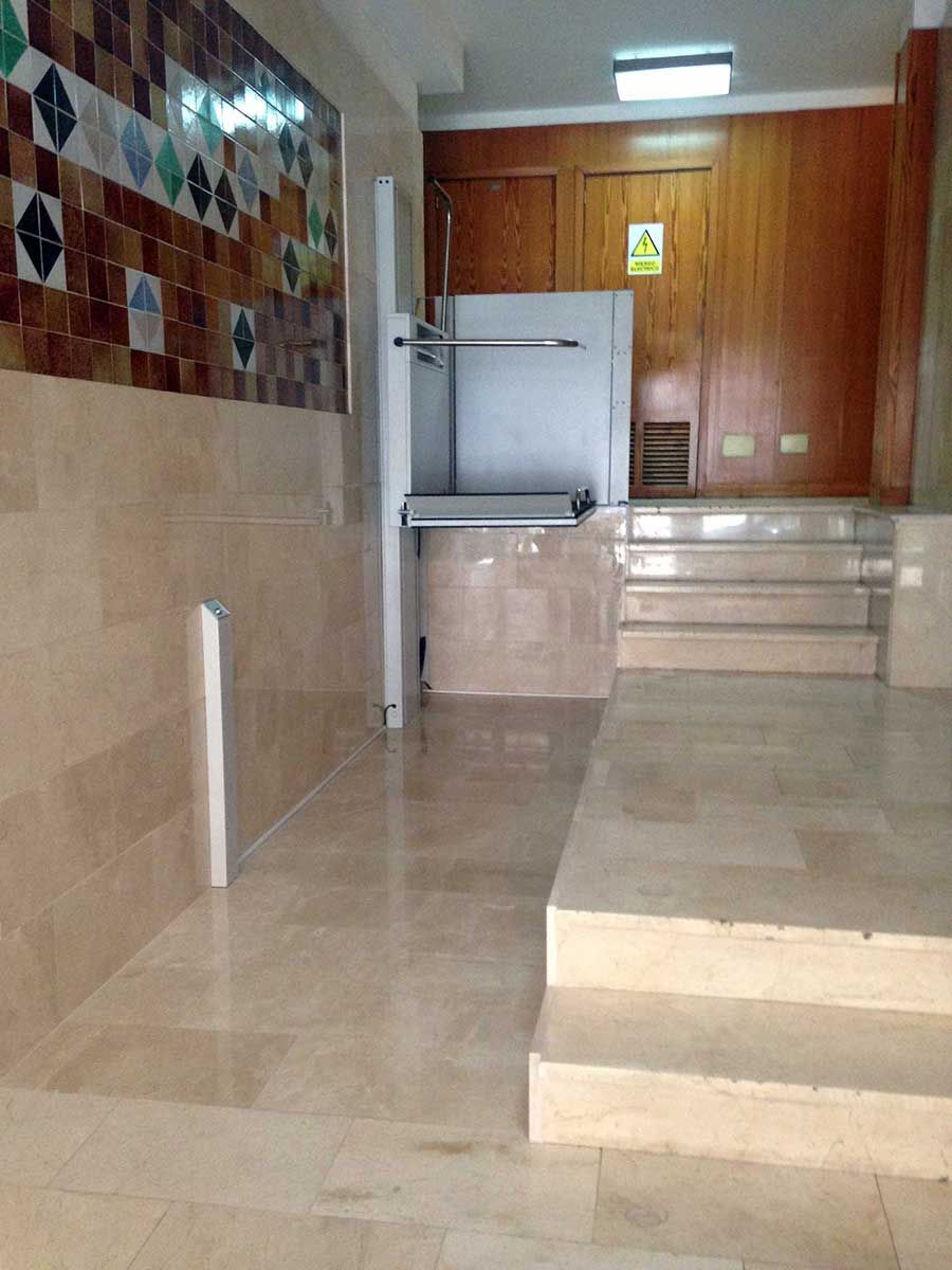 Entrada con plataforma elevadora, escalones y paredes de azulejos en un edificio. Tonos beige y marrón.