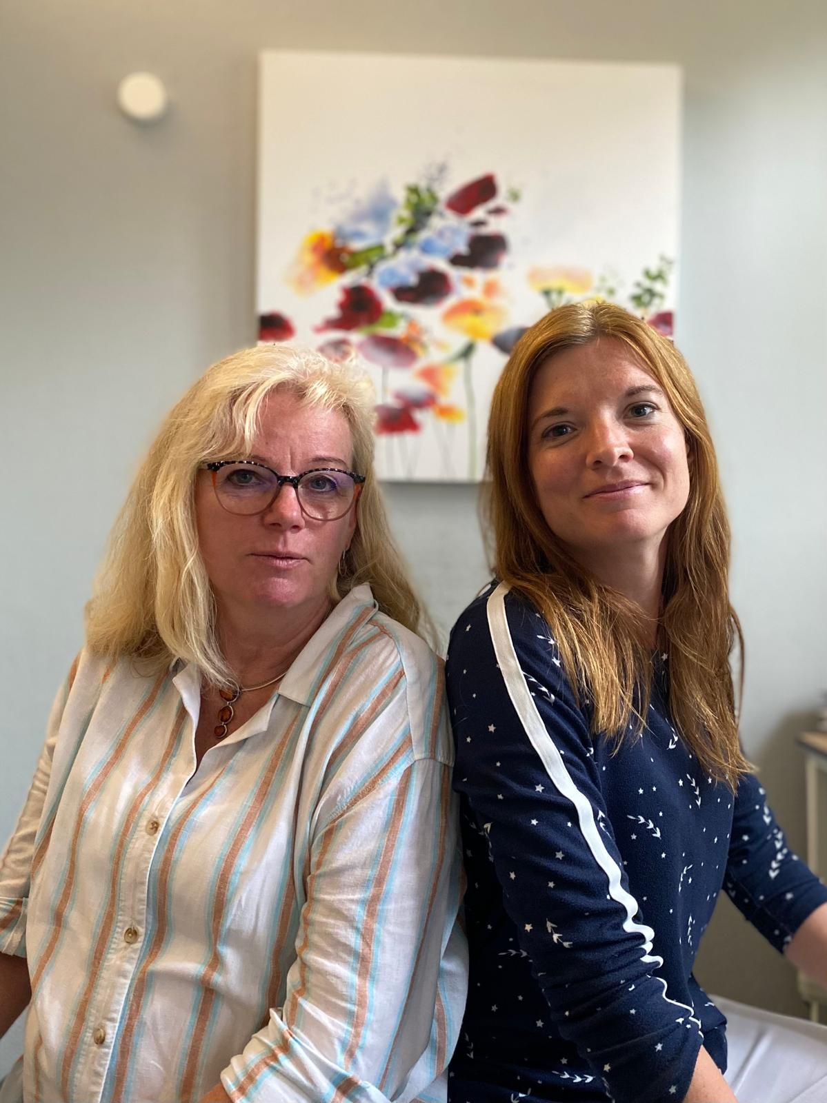 Zwei Frauen, blond und rothaarig, sitzen. Hinter ihnen ein Blumengemälde.