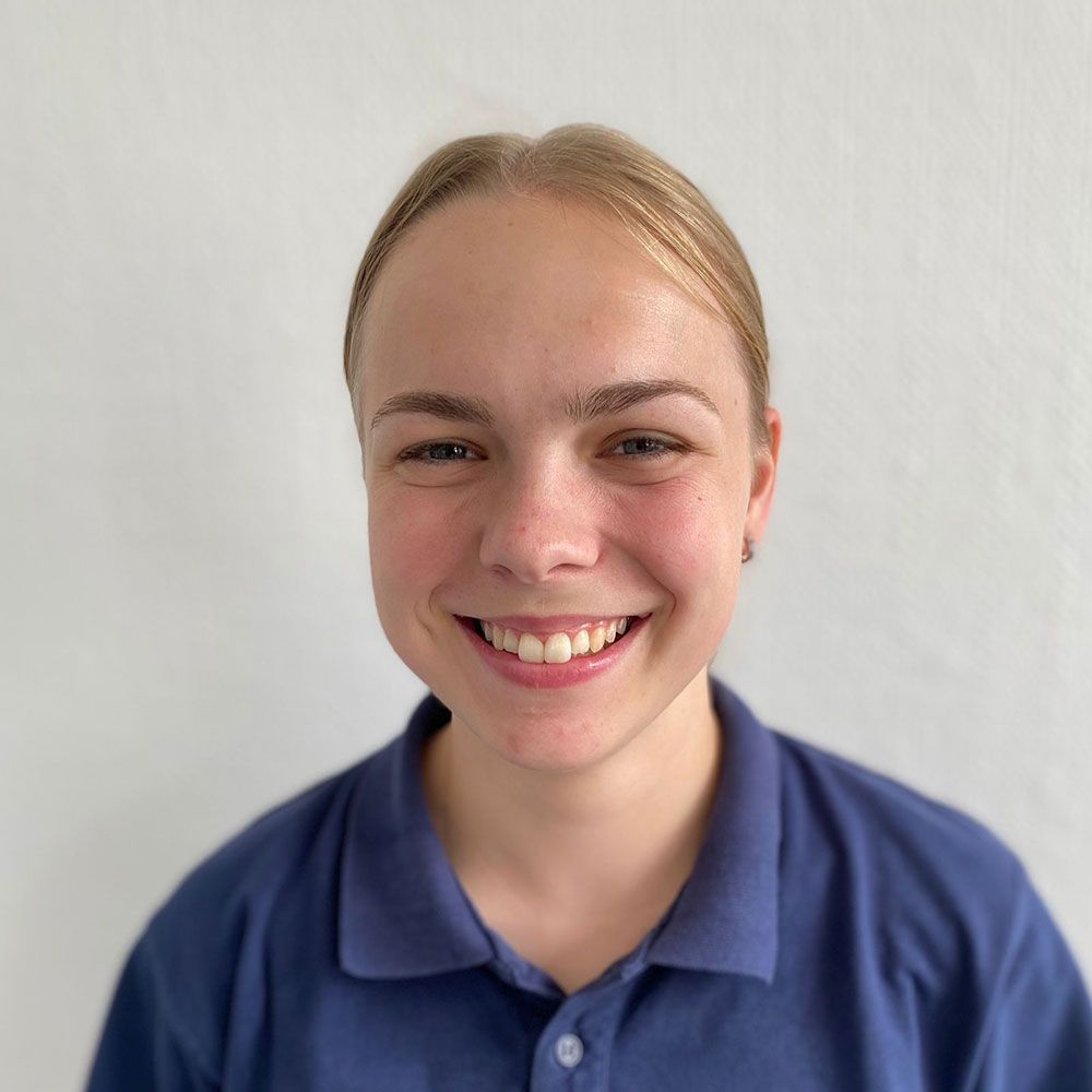 Lächelnde junge Frau in blauem Poloshirt mit zurückgekämmtem blondem Haar vor einer weißen Wand.