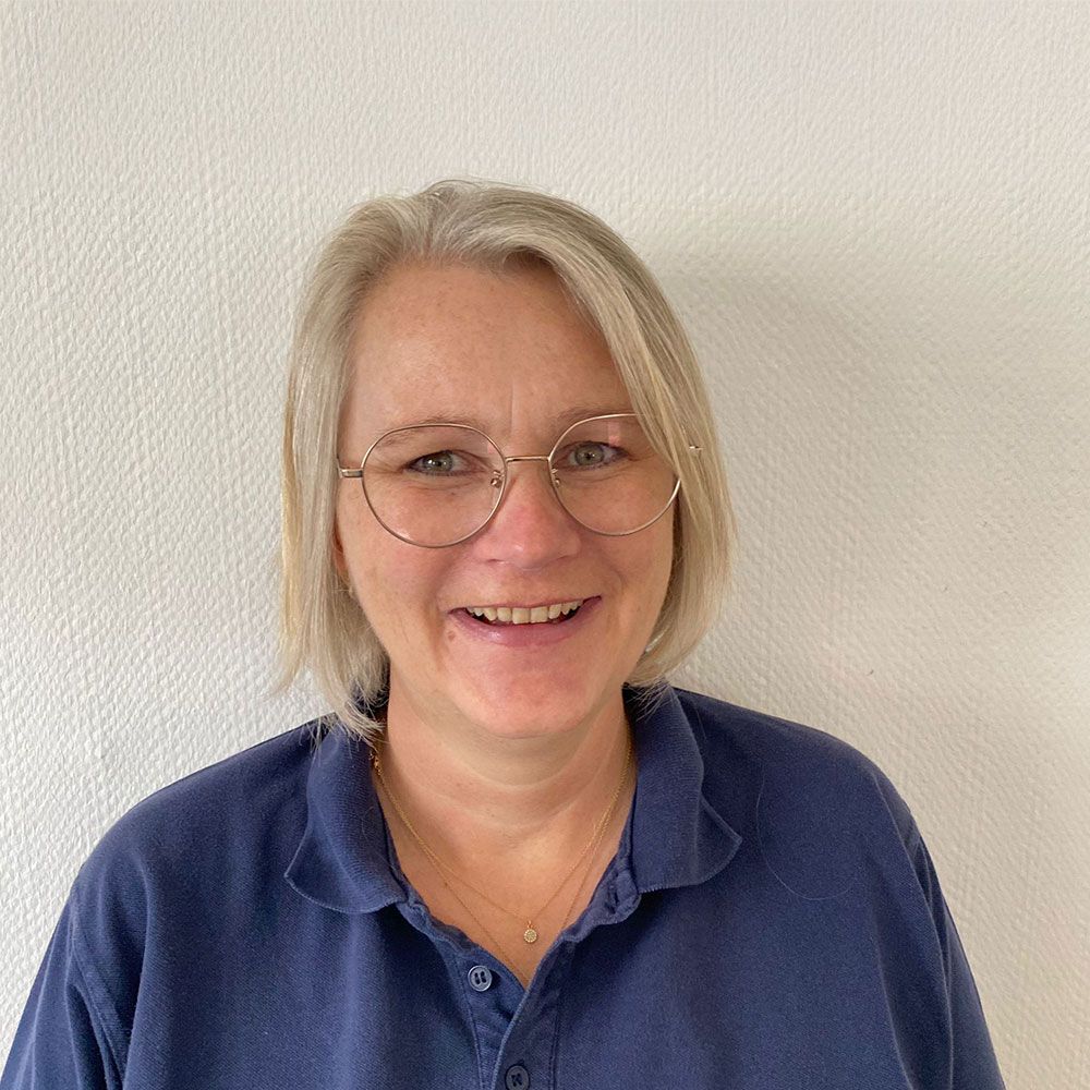Frau mit kurzen, ergrauenden Haaren, Brille und blauem Hemd lächelt vor einer weißen Wand.