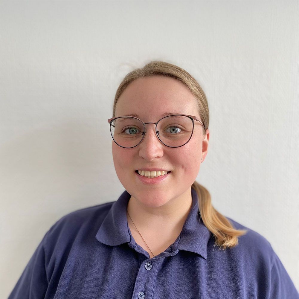Frau mit Brille, lächelnd, trägt ein marineblaues Hemd mit Kragen, blondes Haar zu einem tiefen Pferdeschwanz gebunden.