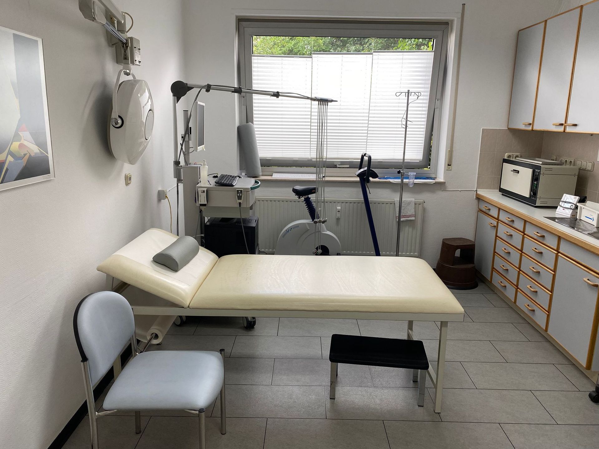 Medizinischer Untersuchungsraum mit Untersuchungstisch, Stuhl, Trainingsgeräten und Schränken.