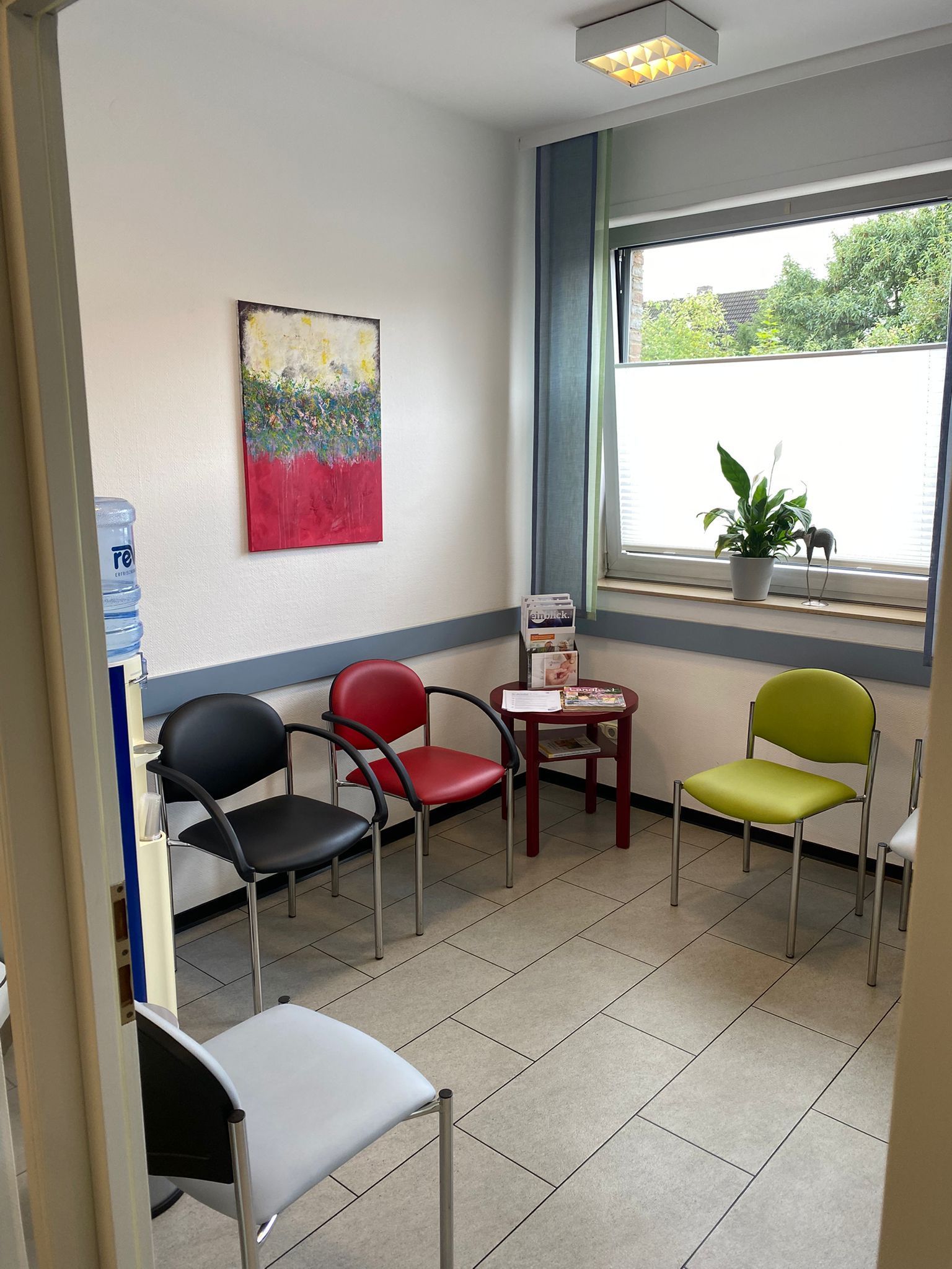Wartezimmer mit Stühlen in verschiedenen Farben, einem Gemälde und einem Fenster.
