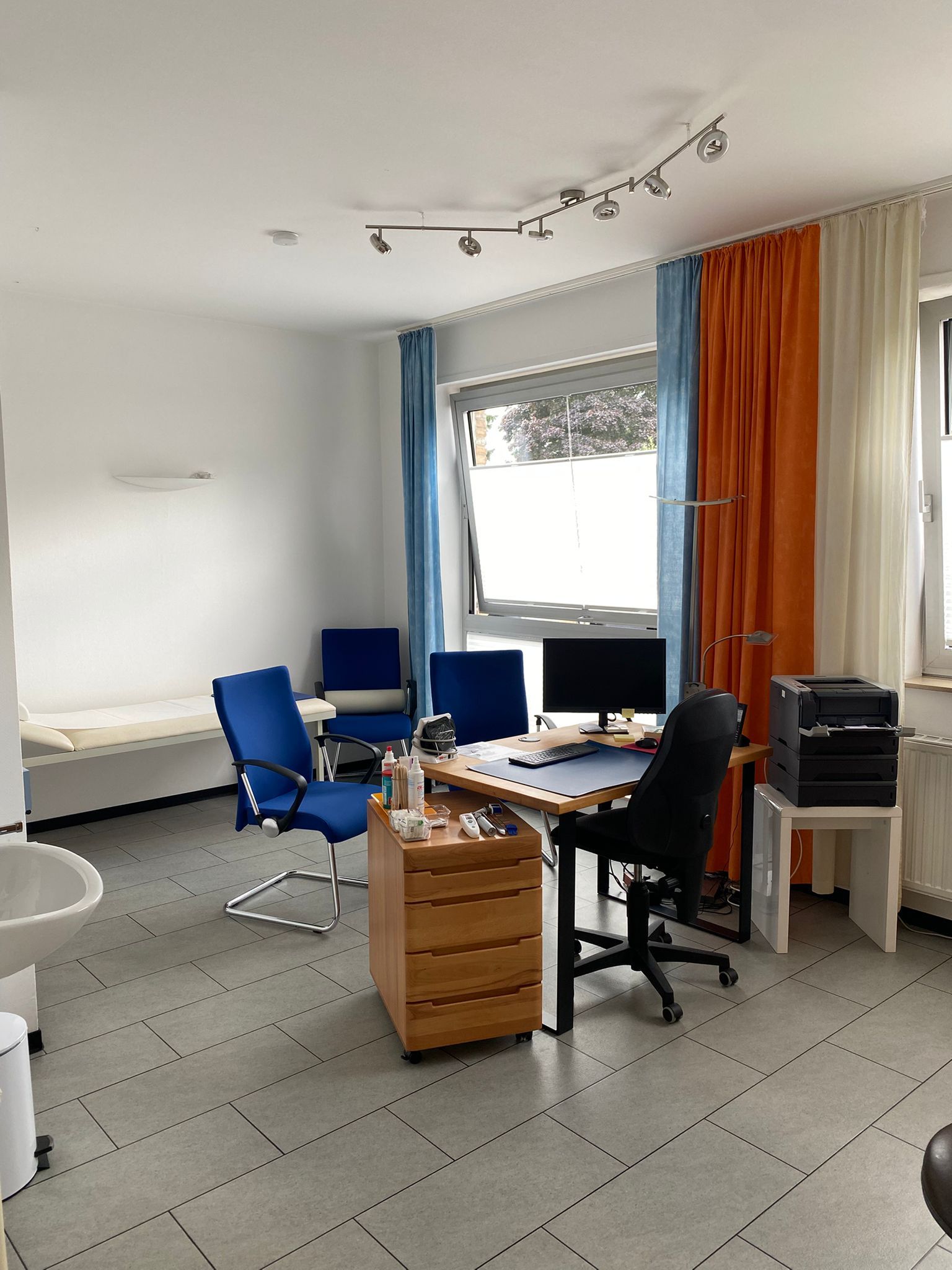Büro mit Schreibtisch, blauen Stühlen, Fenster mit bunten Vorhängen und einem Drucker.