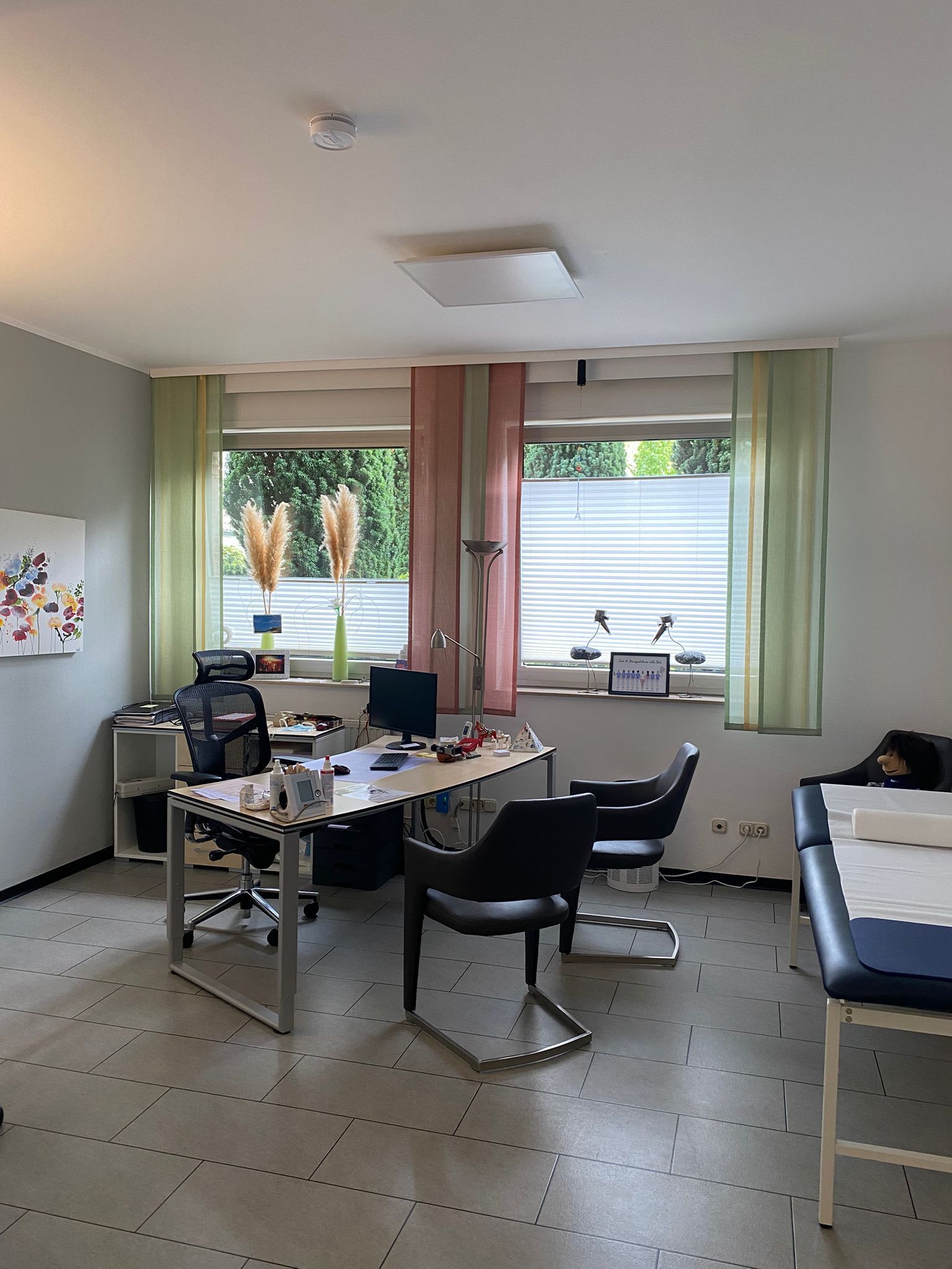 Innenraum einer Arztpraxis mit Schreibtisch, Stühlen und Untersuchungstisch. Grüne und rosa Vorhänge verdecken die Fenster.