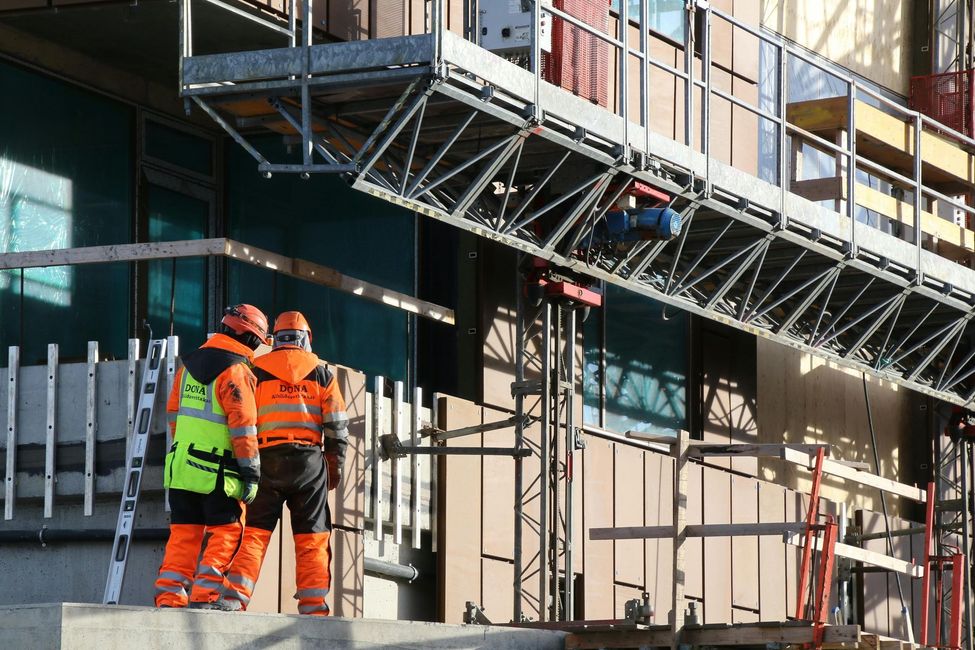 Obreros de la construcción con chalecos naranjas miran hacia los andamios en la fachada de un edificio.