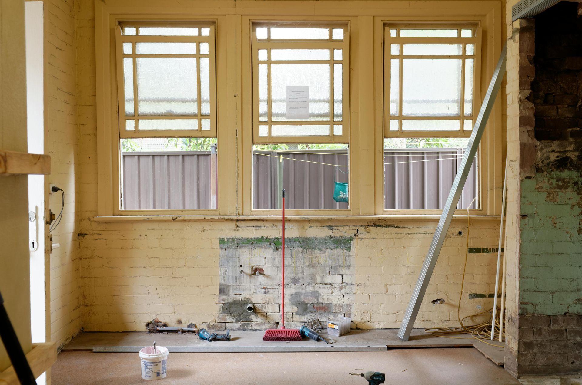 Renovación de habitación interior con tres ventanas, ladrillo visto, herramientas de construcción y una escoba roja.